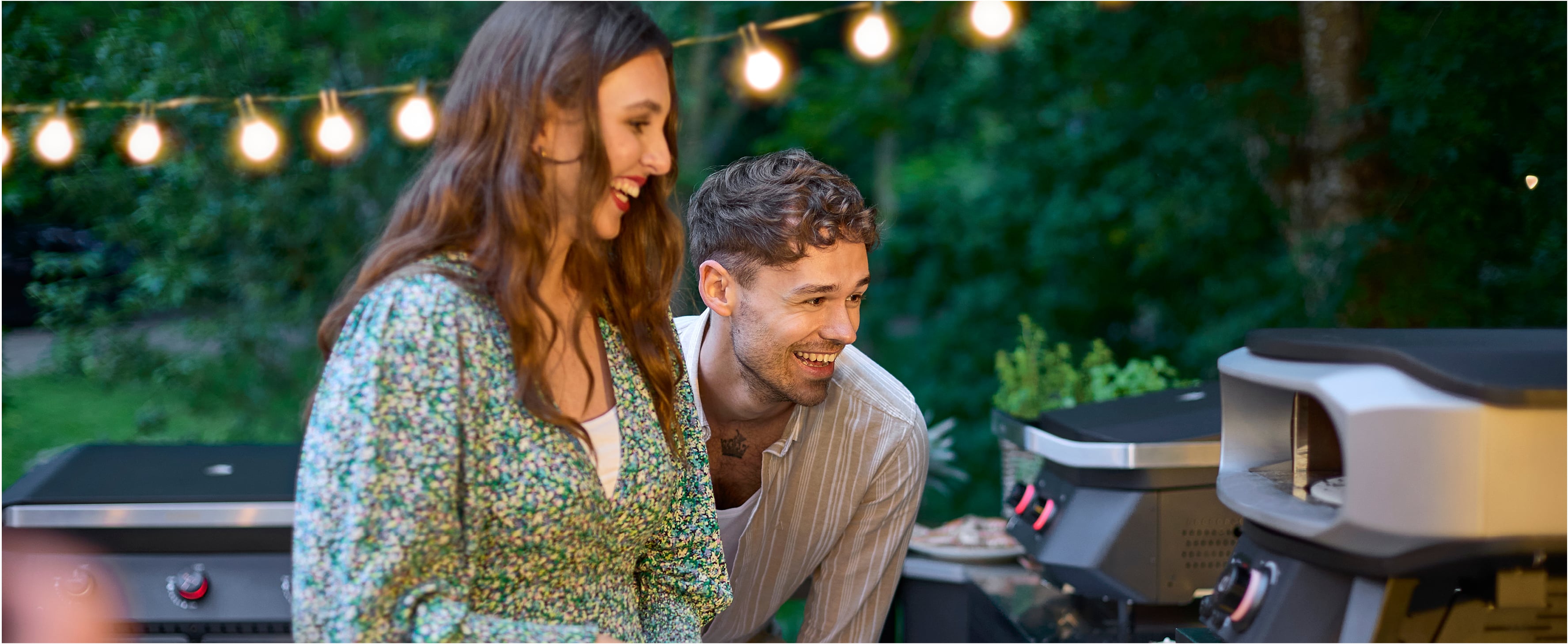 Lächelndes Paar genießt Sommerabend am Cozze Pizzaofen und Gasgrill im Garten – gemütliche Atmosphäre mit Lichterketten