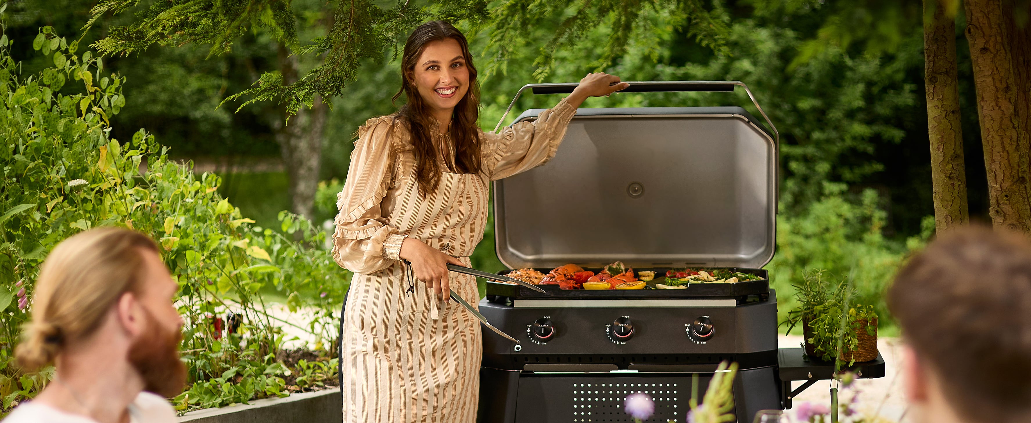 SLächelnde Frau in Schürze grillt buntes Gemüse auf Cozze Gasgrill mit 3 Brennern im Garten – Sommerstimmung mit Freunden