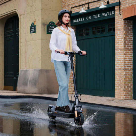 Frau mit Helm fährt Navee GT E-Scooter durch Pfütze auf Kopfsteinpflaster – spritzwassergeschützt und stadttauglich